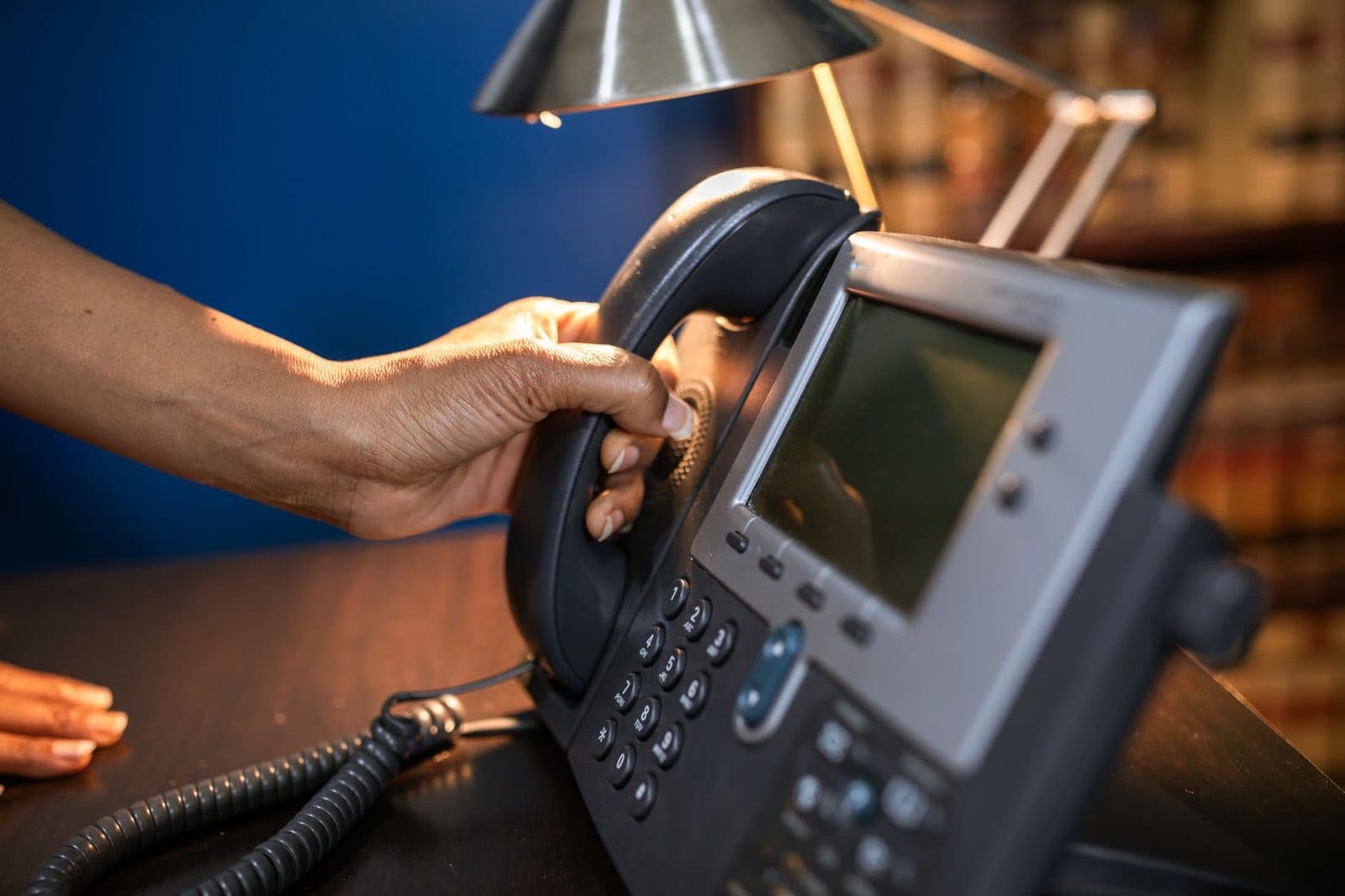 A hand reaching for the handset of an office desk phone.