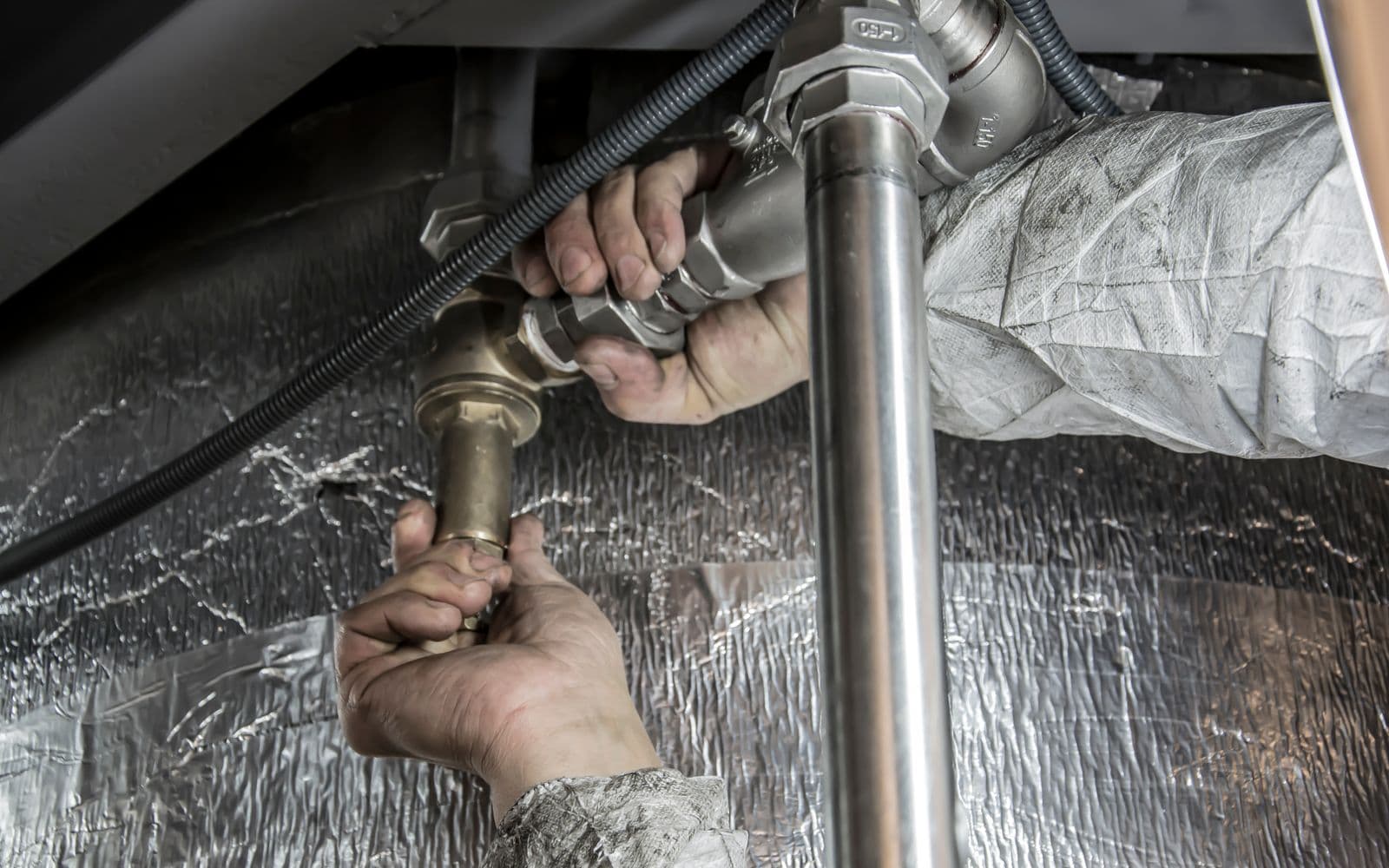 A plumber's hands tightening a brass fitting on the supply pipes under a sink.