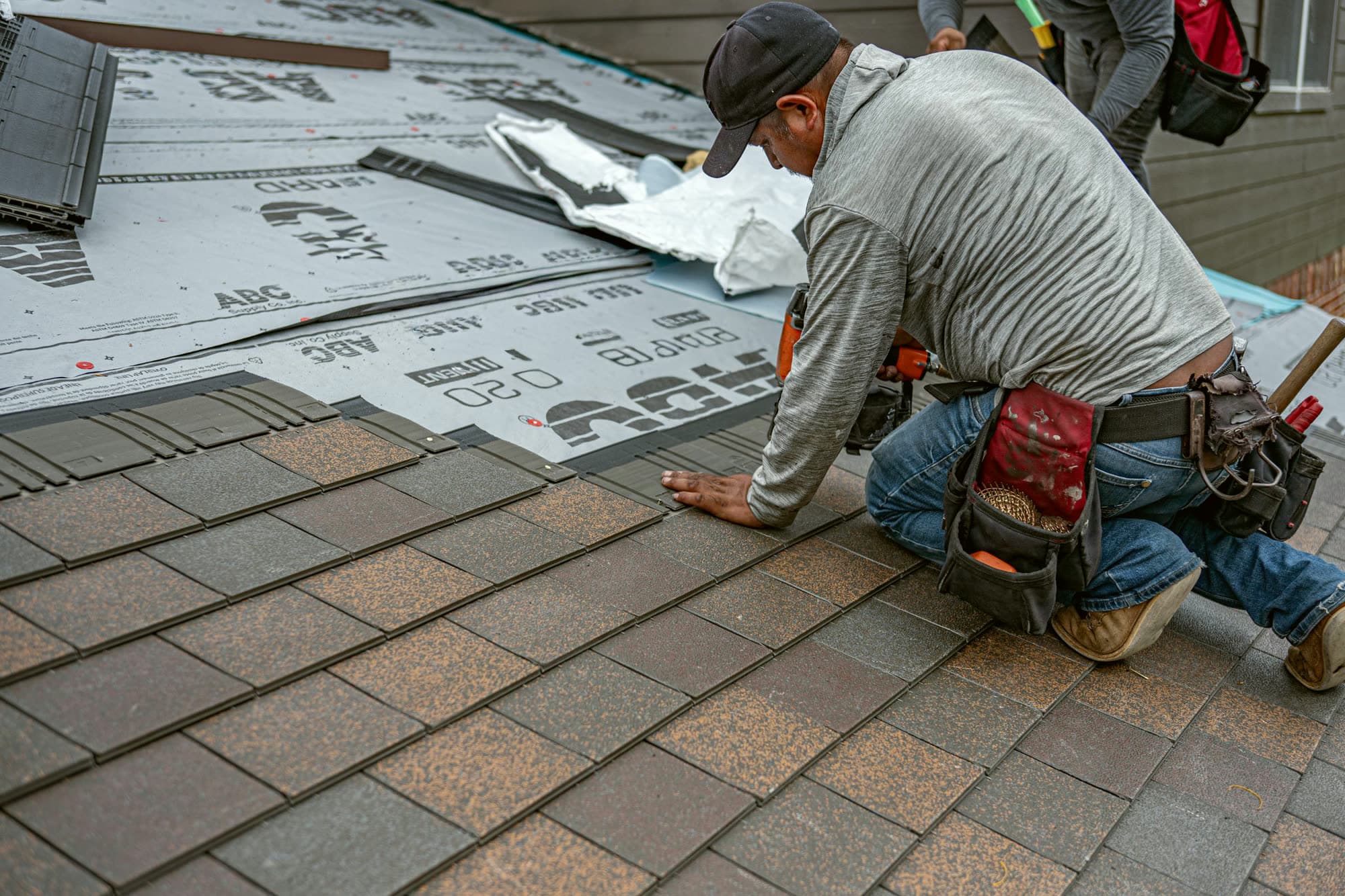 A roofer with a tool belt kneeling to lay shingles.